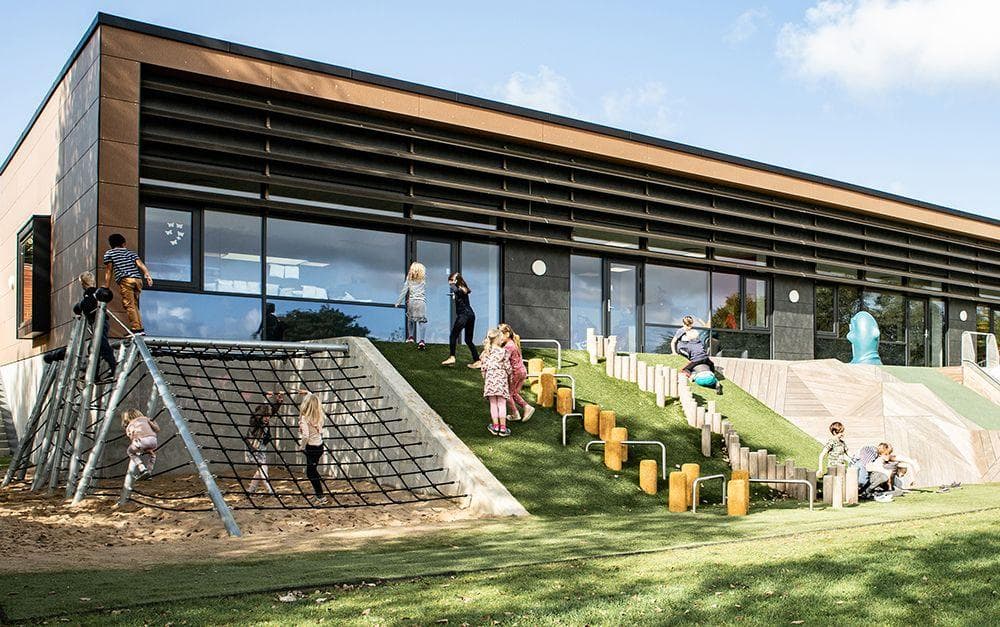 Moderne pavillon med træ- og betonfacade, børn leger på skrå legeredskab og gule klodser på grønt underlag