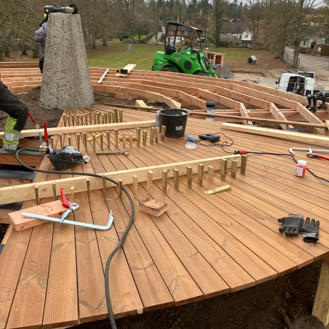Træbygning under konstruktion med planker, værktøj og grøn gravemaskine på stedet