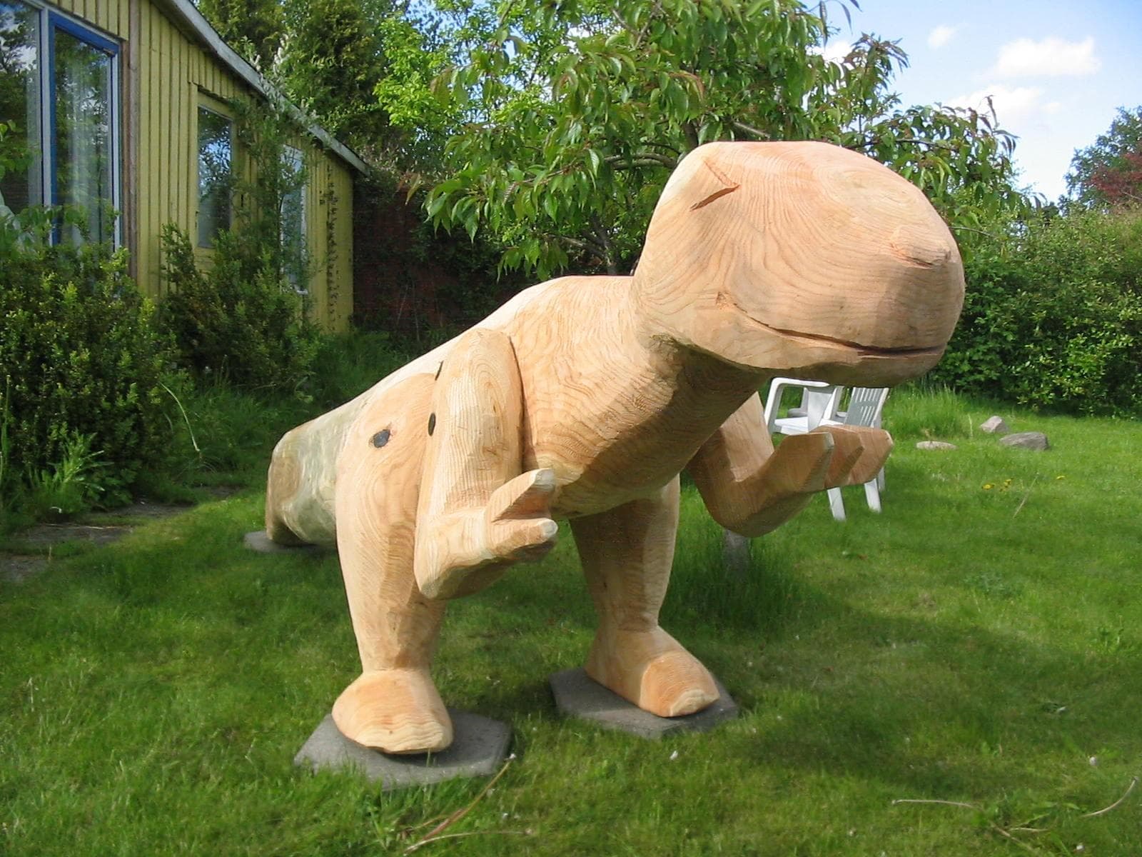 Træskulptur af dinosaurus på græsplæne foran drivhus og grønne buske