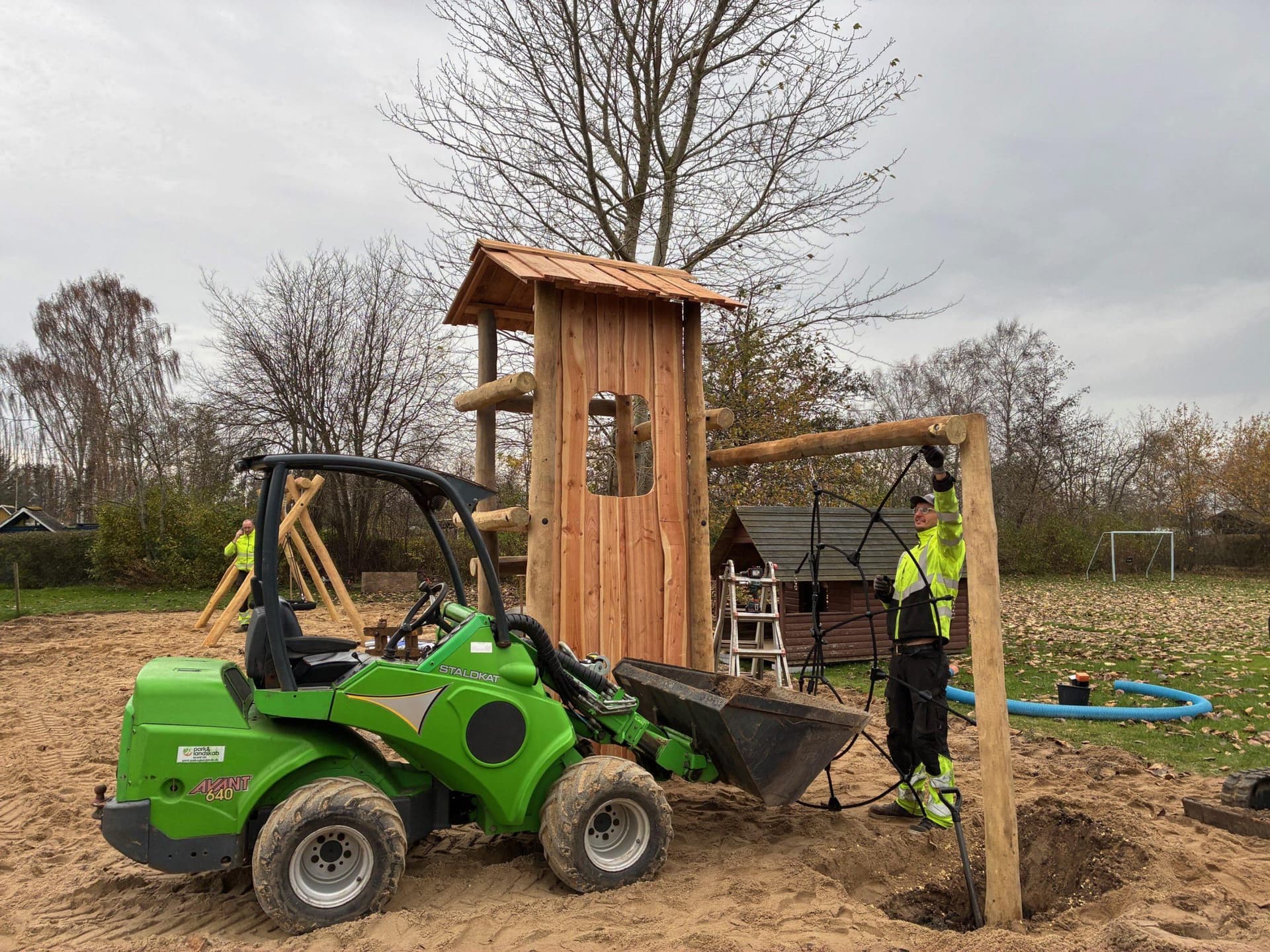 Grøn minigraver ved træhutte i træ og voksen i arbejdstøj på sandflade