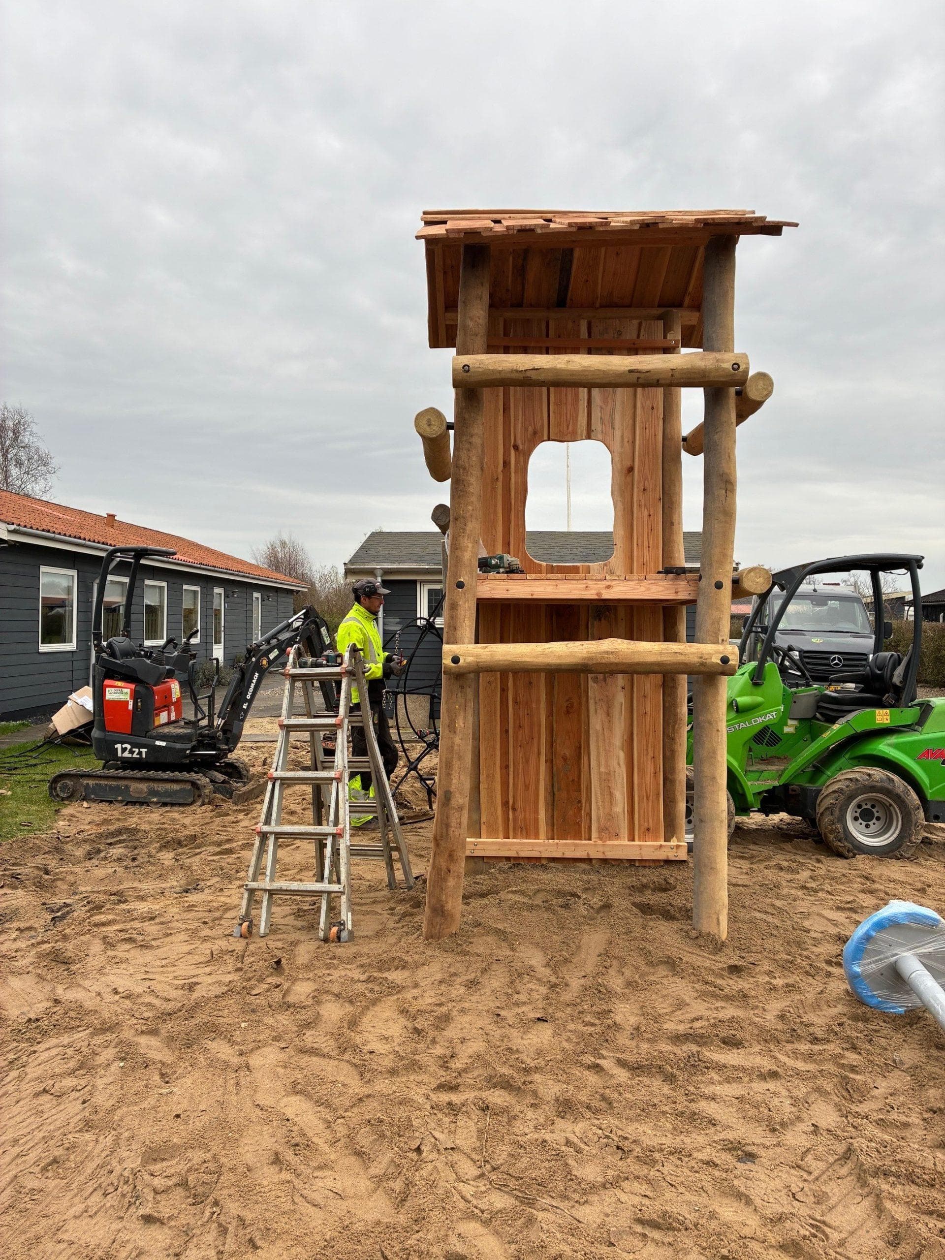 Træ-legetårn under konstruktion på sandunderlag med arbejdere