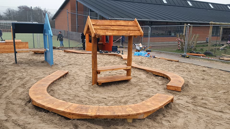 Træhytte med rødt tag på sandunderlag med buet træbænk omkring