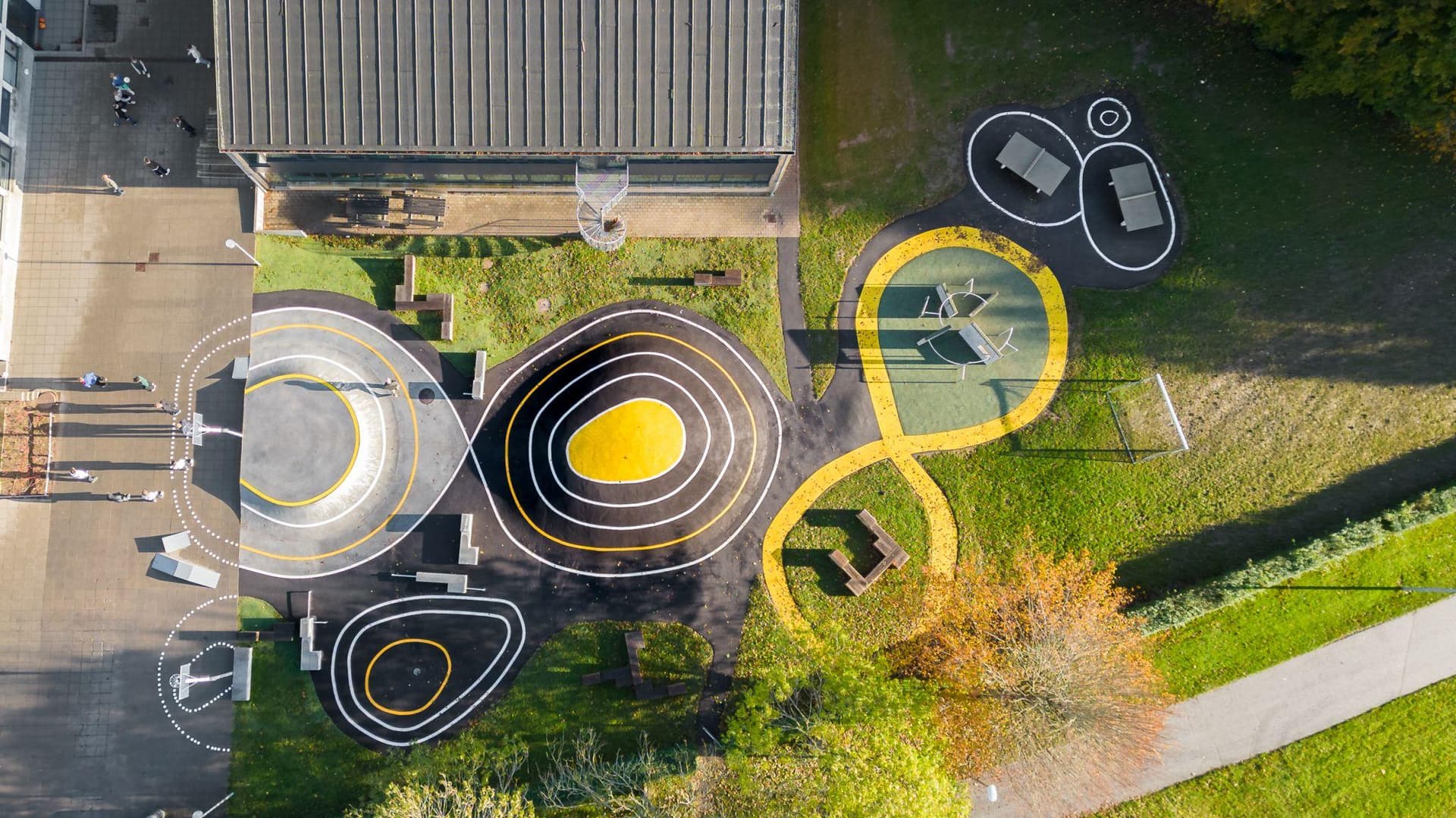 Fugleperspektiv af legeområde med spiralformede rutsjebaner i gul og sort, græsunderlag og bygning