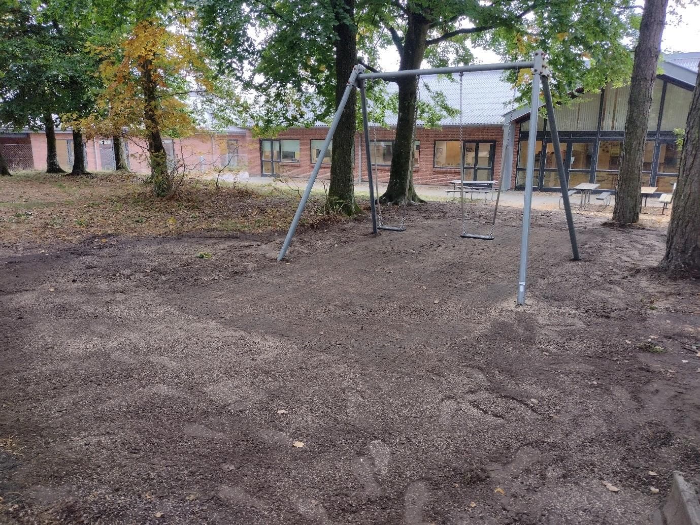 Legeområde med gynger på jordunderlag blandt høje træer ved mursten-skolebygning