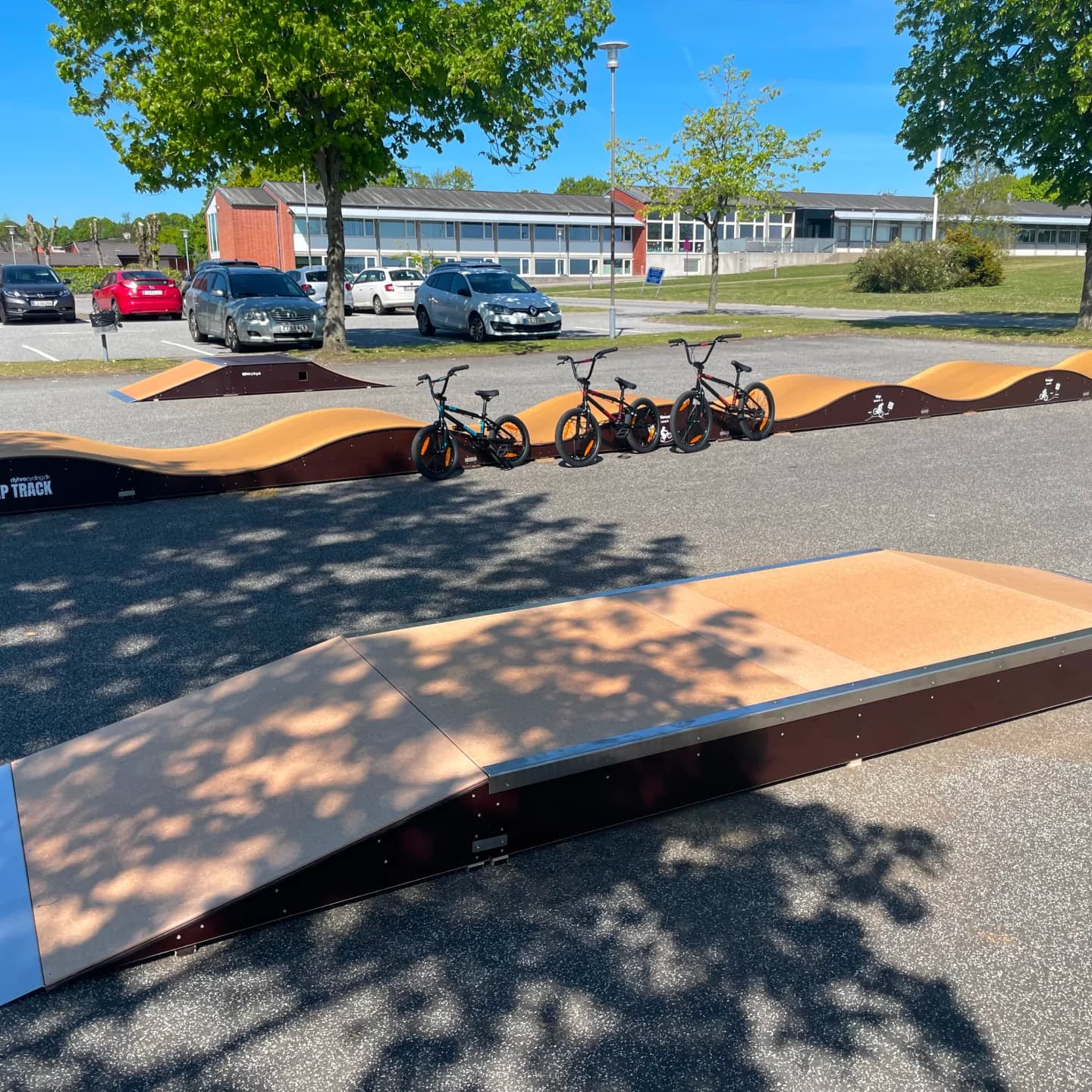 Pumptrack i orange og blå med tre børnecykler, parkeringsplads og bygninger i baggrund