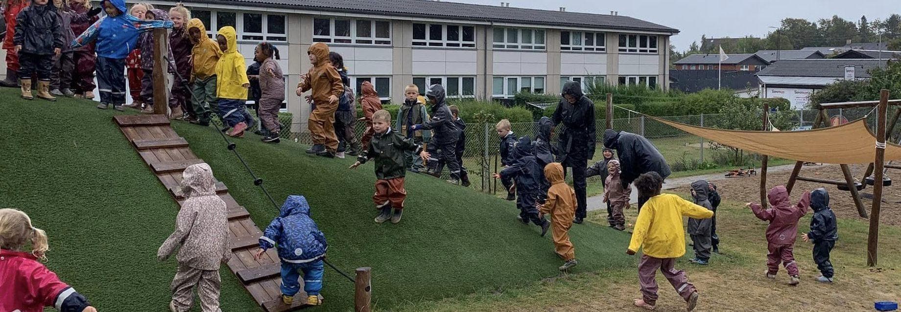 Børn og voksne på kunstgræsplæne med legestrukturer og institutionsbygning i baggrunden