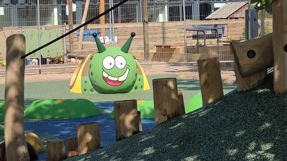 Grøn rutsjebane med smilende figur blandt træstammer på gummifliser