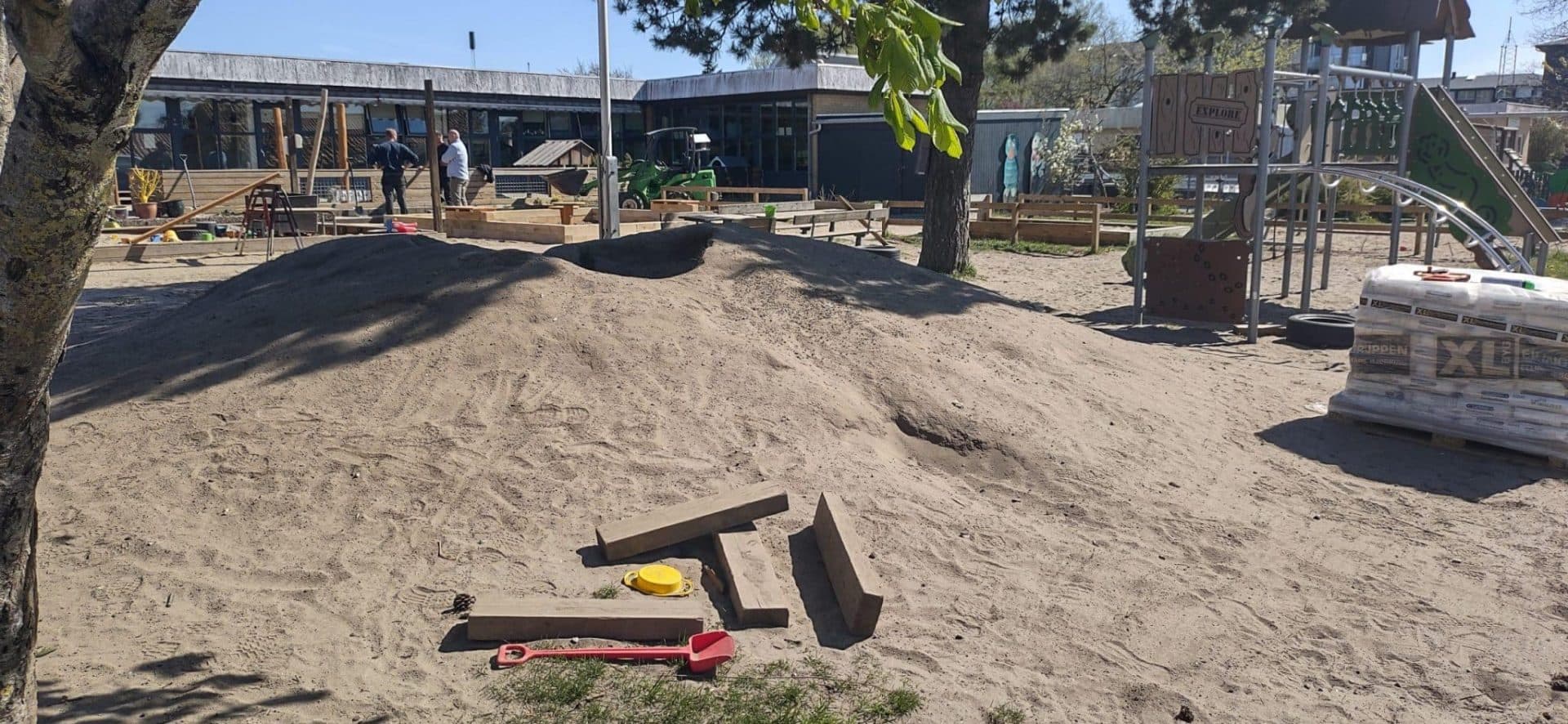 Sandspilareal med udgravningsudstyr, træer og bygninger i baggrunden