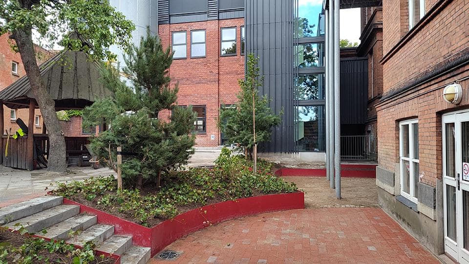 Rødt plantebed med buske og træer på rødt flisunderlag mellem teglstensbyggeri