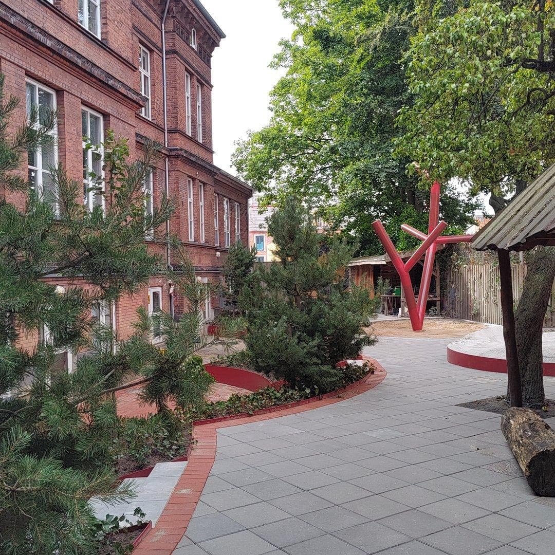 Rødt kunstværk blandt træer på terrassebelægning foran rødt mursten-skolebygning