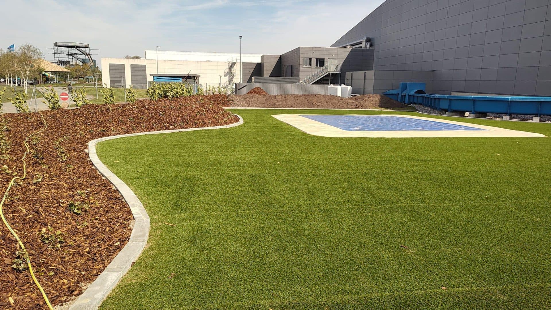 Kunstgræsområde med hvid afgrænsning, mulch-bed og blå springmåtte ved industriel bygning