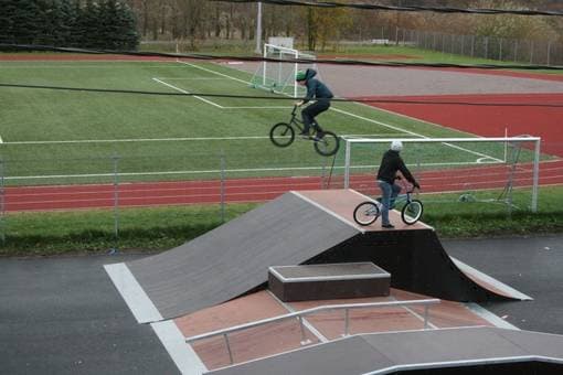 Børn på BMX-cykler på sort asfaltramp med kunstgræsbane i baggrunden