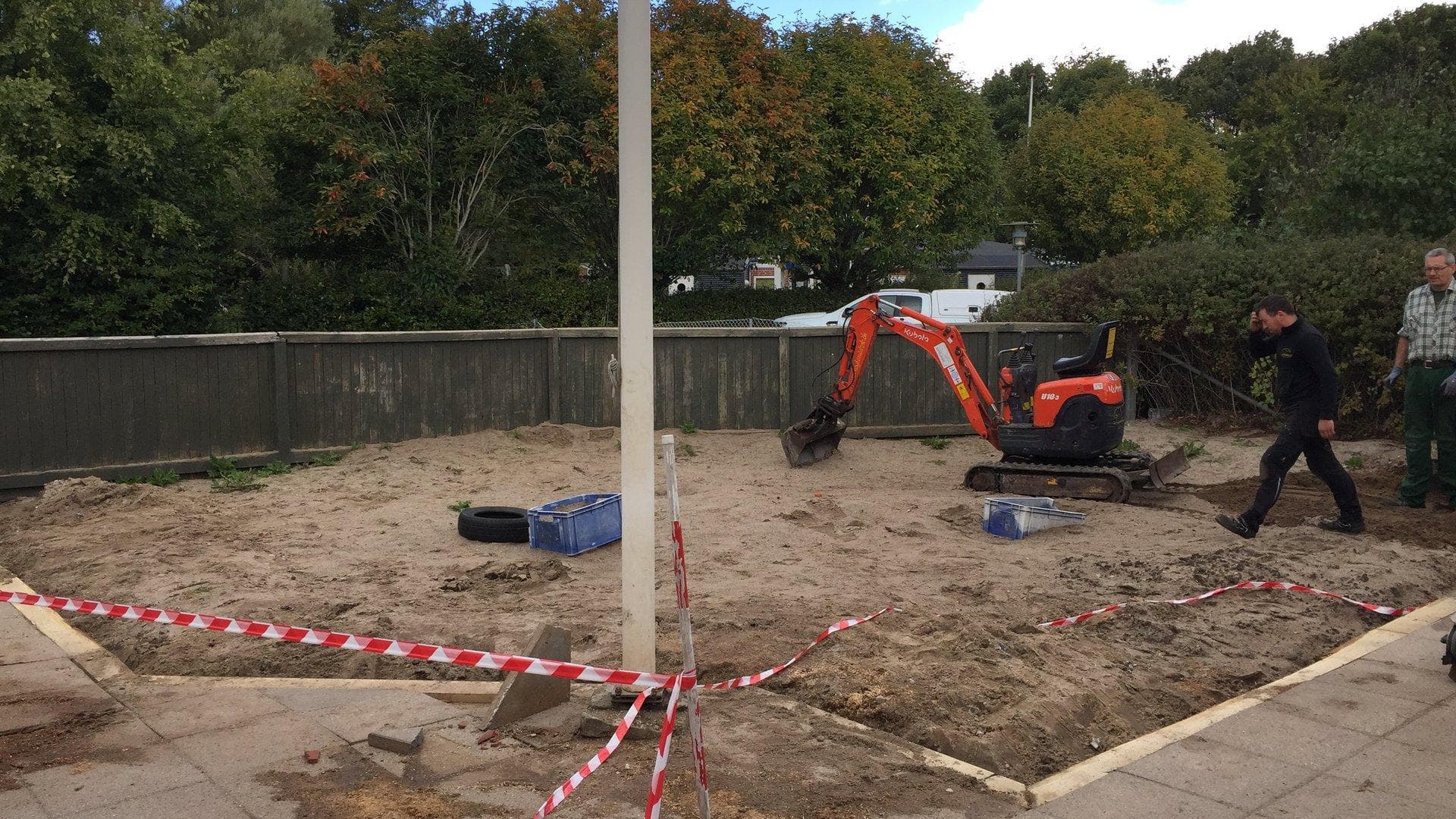 Sandkasse under anlæggelse med rød minigræver og arbejdere på område omgivet af rødt-hvidt afspærringsbånd