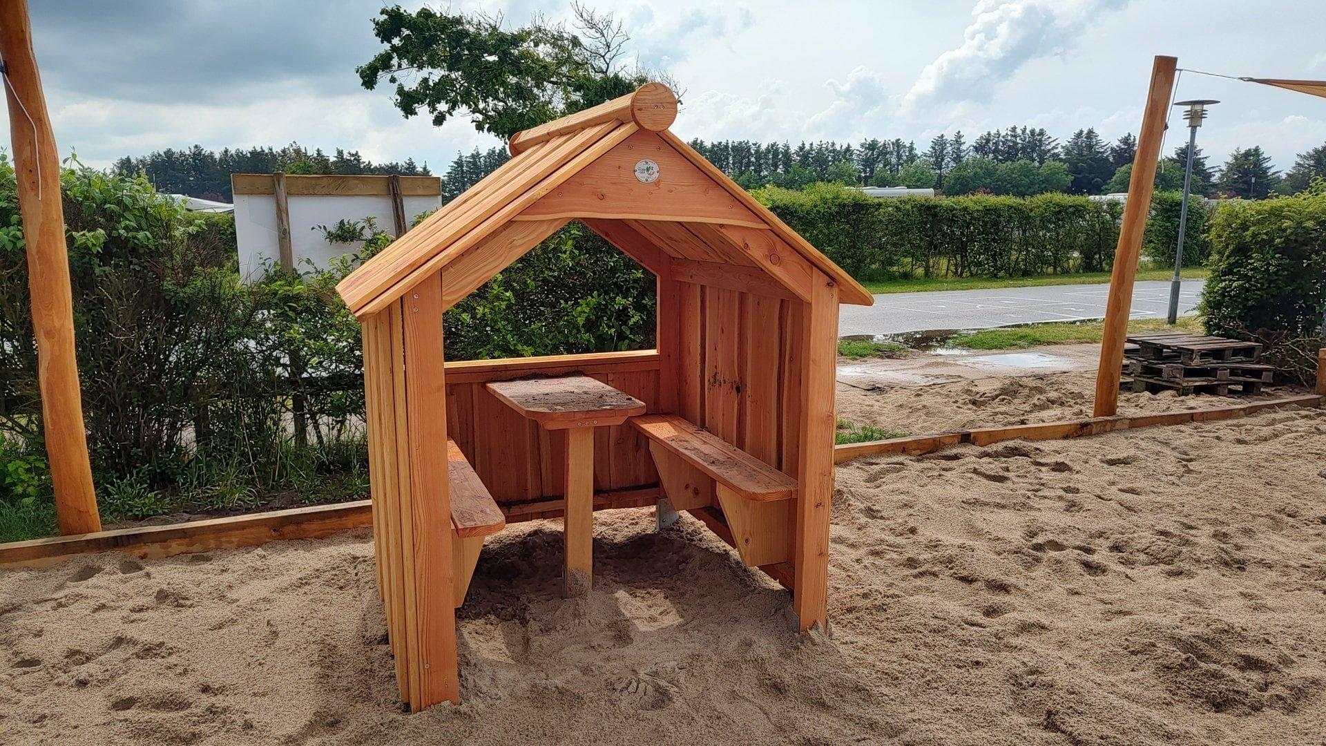 Træhytte med bænk indeni på sandflade, omgivet af grønne områder og træer