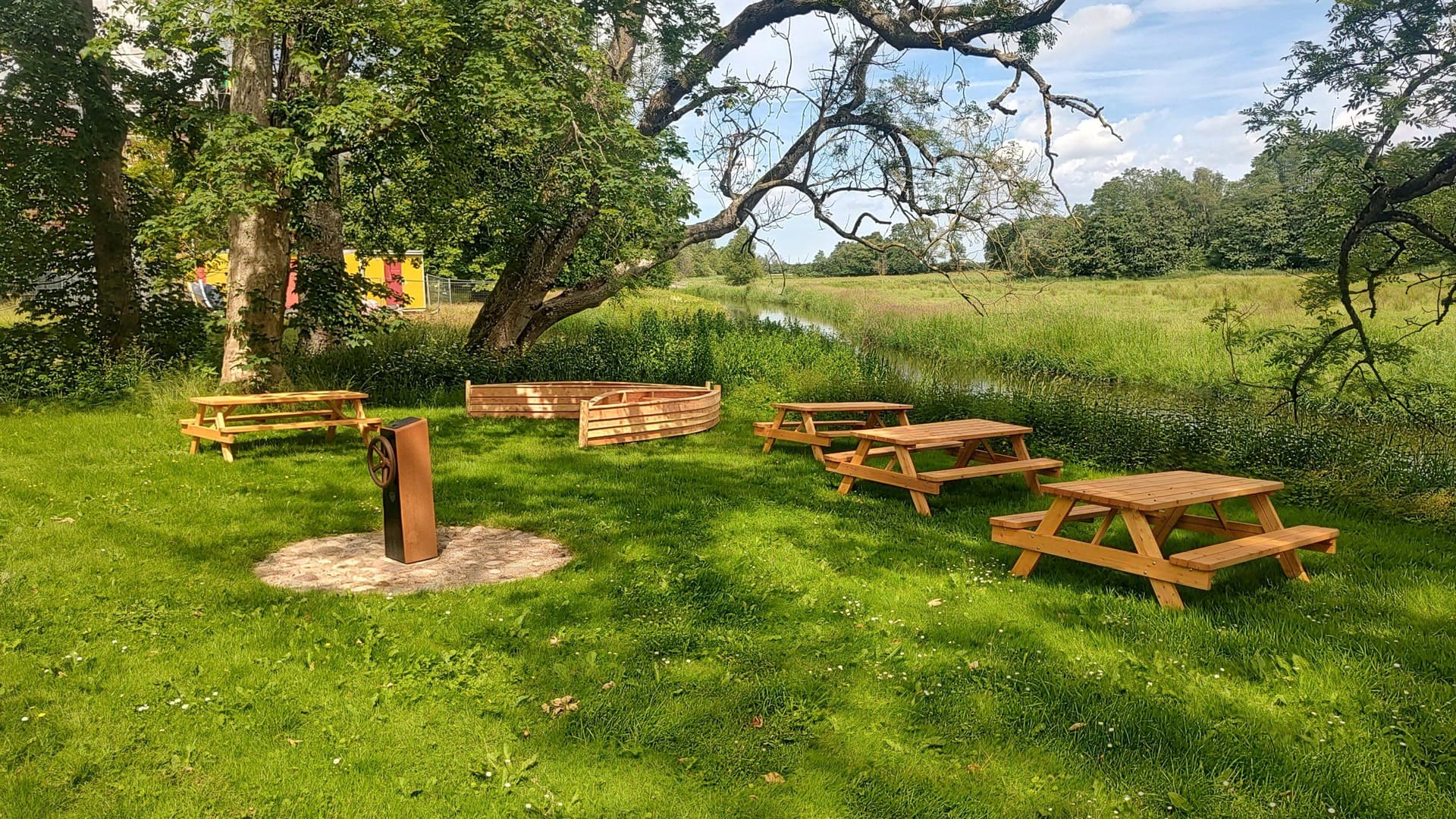 Træpiknikborde og bålsted på grønt græsareal under gamle træer ved vandbred