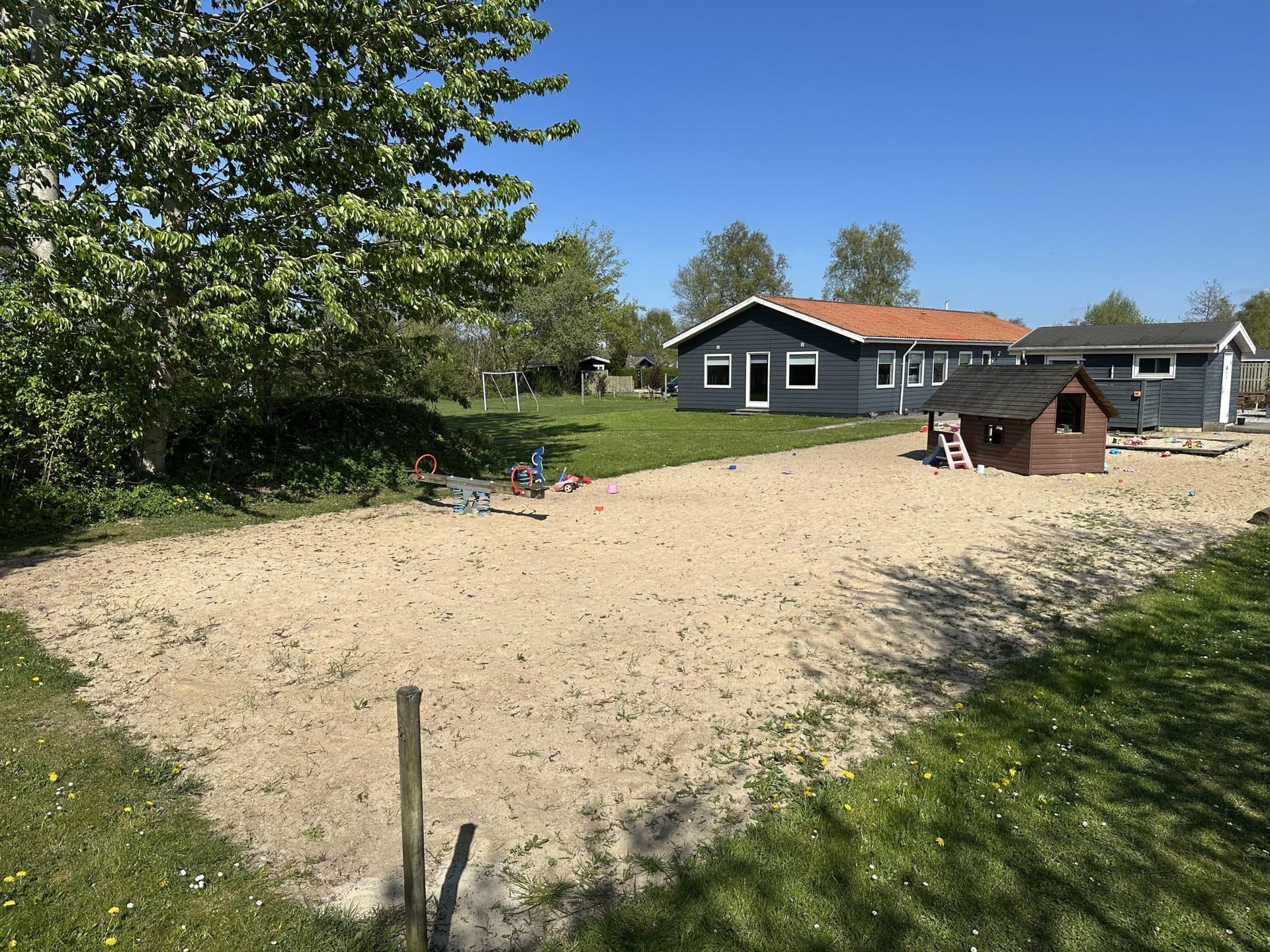 Sandlegeplads med træhytter, græsarealer og løvtræer i baggrund