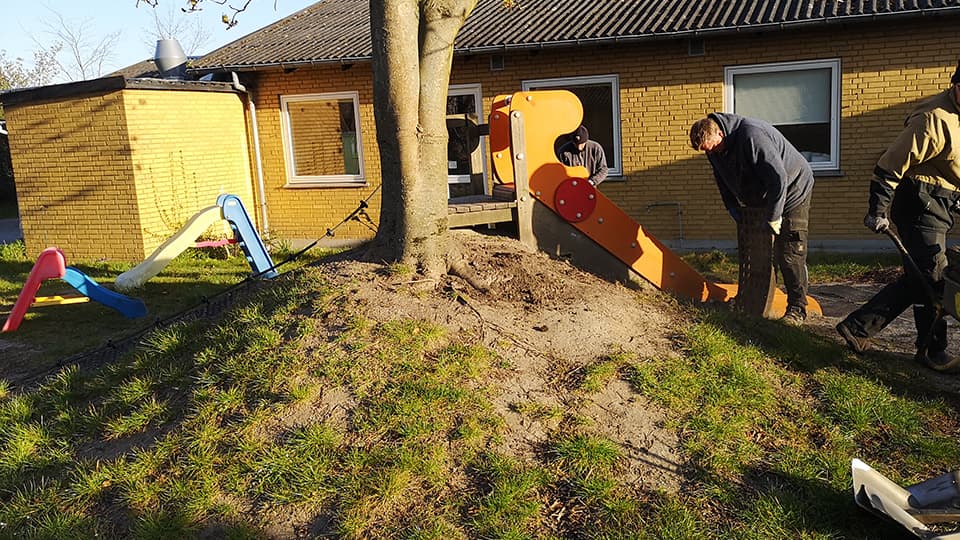 Børn og voksne arbejder med jordarbejde på skolegård ved gult murerstenhus