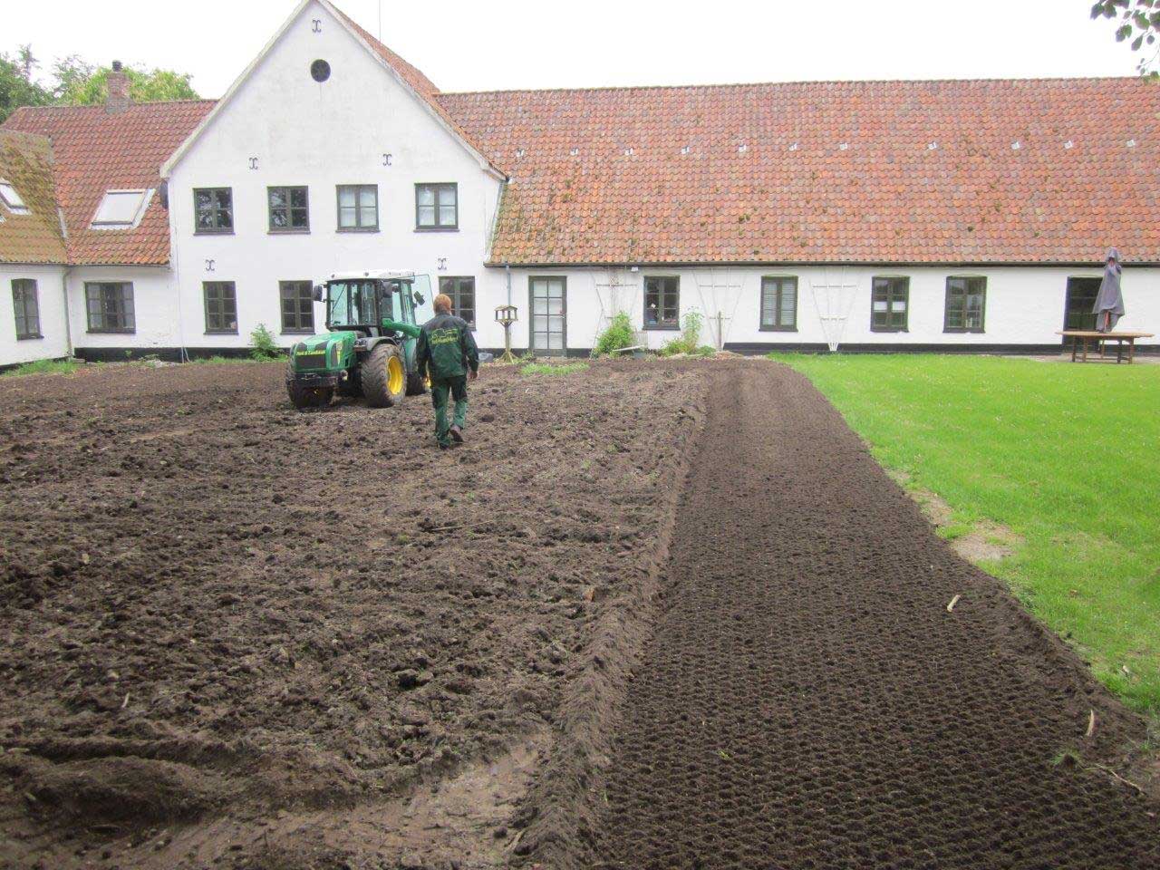 Traktor forbereder jordplots ved større brick-bygning med grønt område