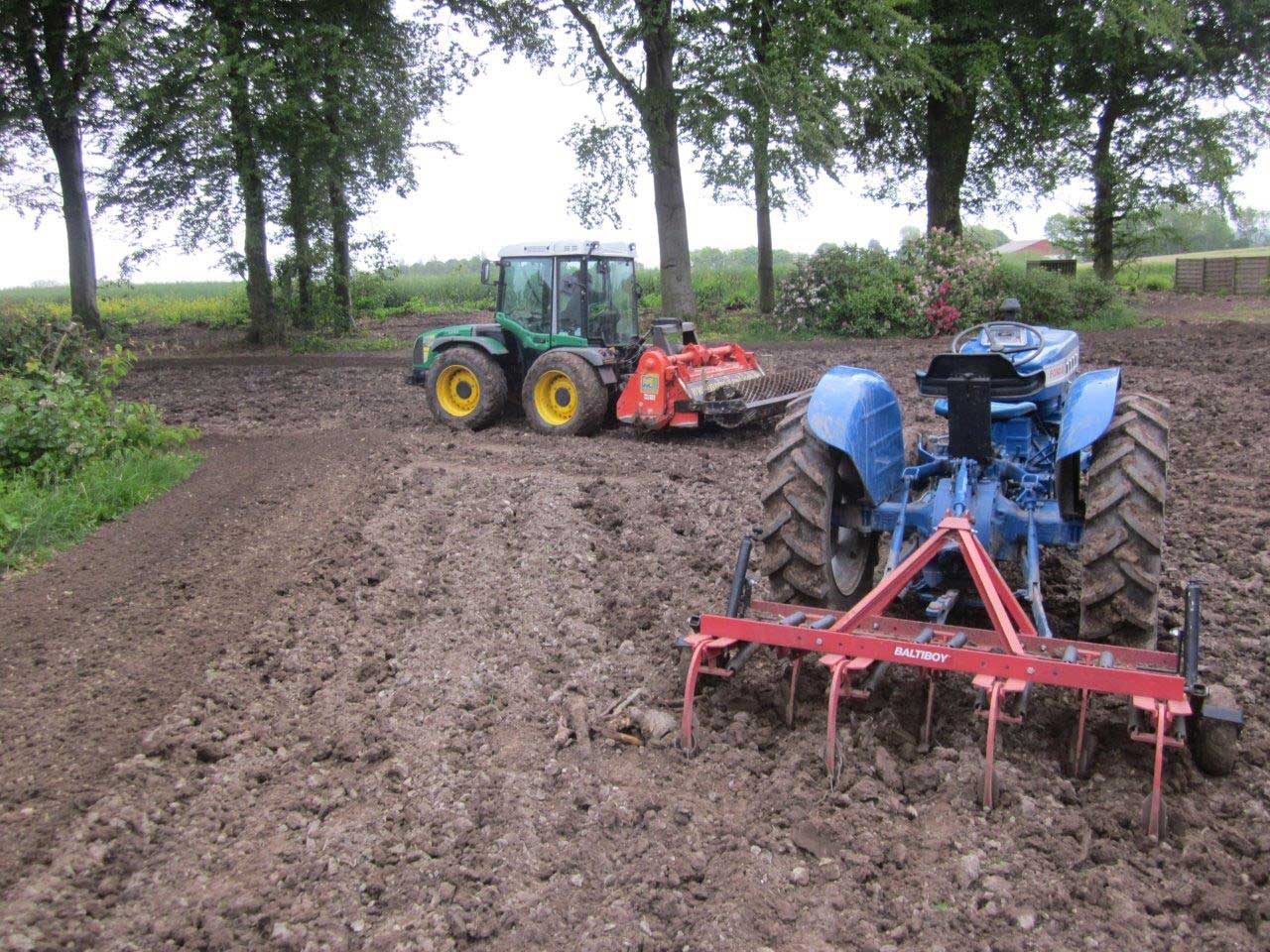 Blå traktor med rød harve på pløjet mark med gul traktor i baggrunden