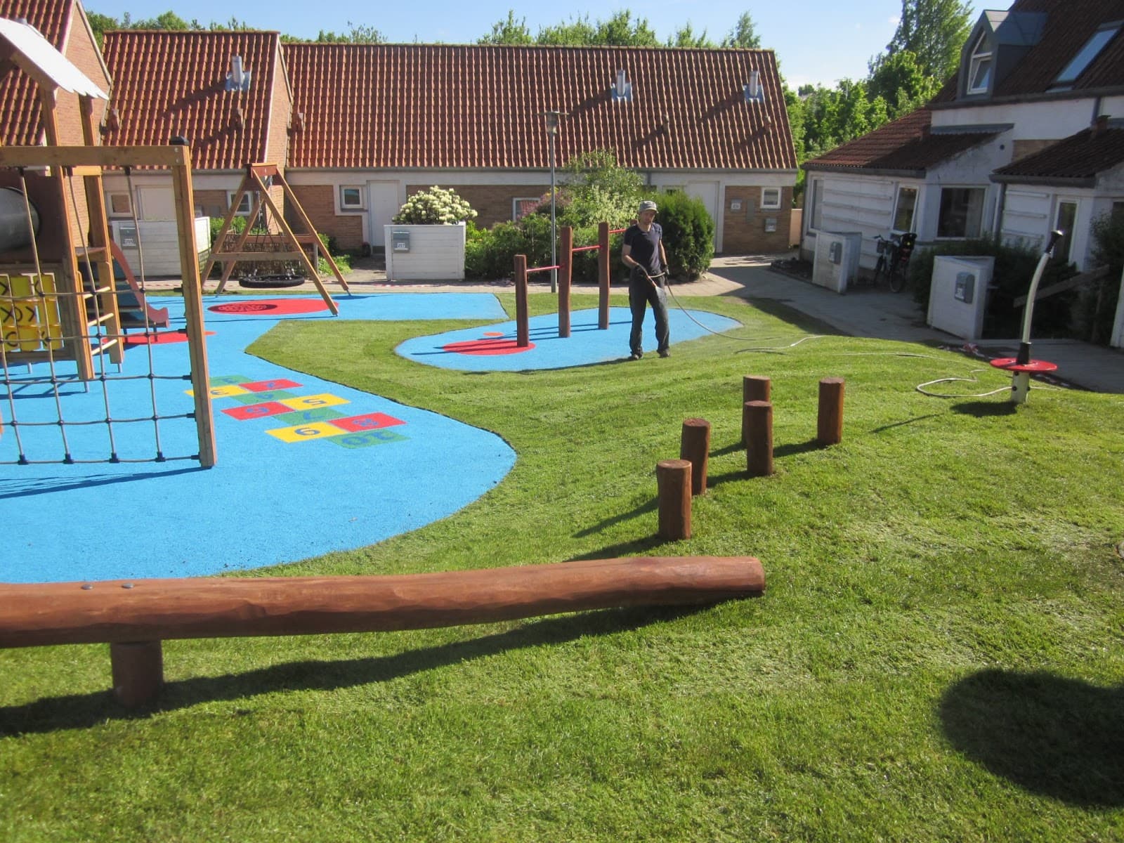Legeområde med blåt gummifaldunderlag, træredskaber og grønt kunstgræs ved beboelsesejendom