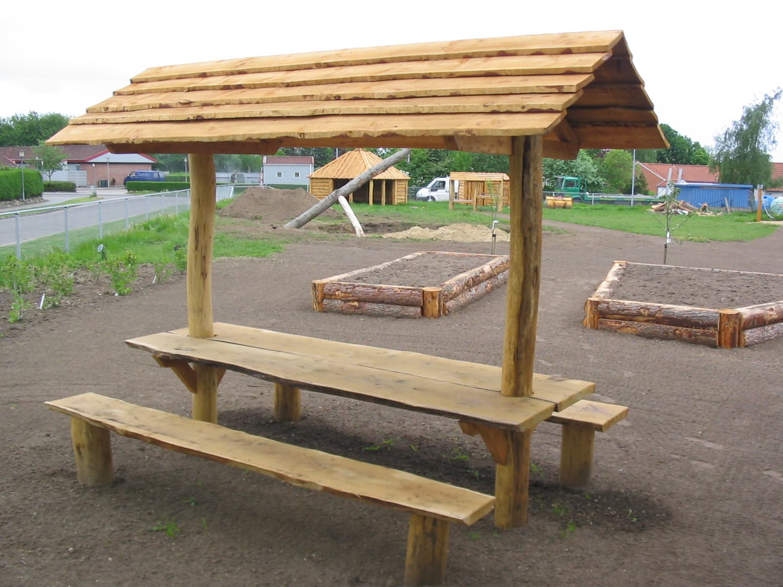 Træpavillon med bænke på asfaltkryds med sandkasser og legeredskaber i baggrund