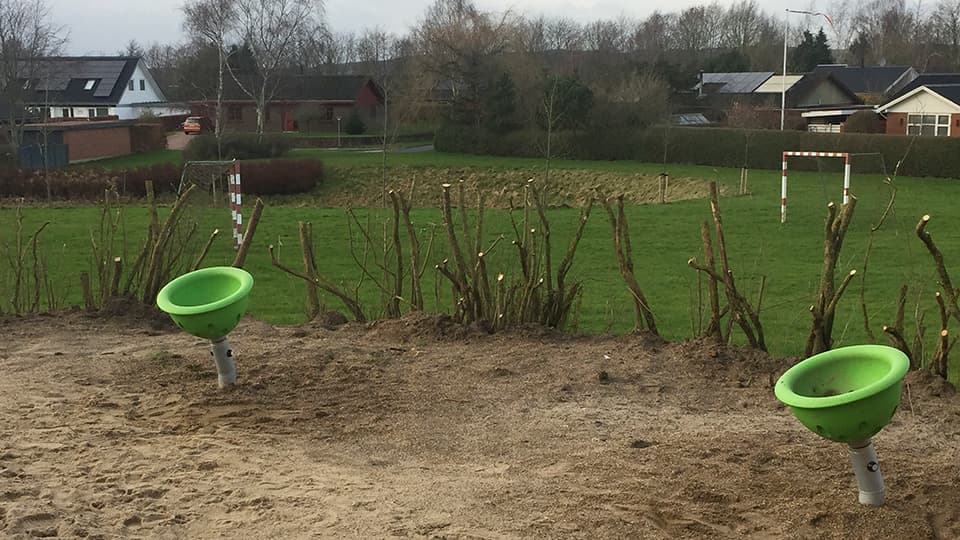 To grønne trætopfodringskåle på sandunderlag blandt pæle med boligområde bag