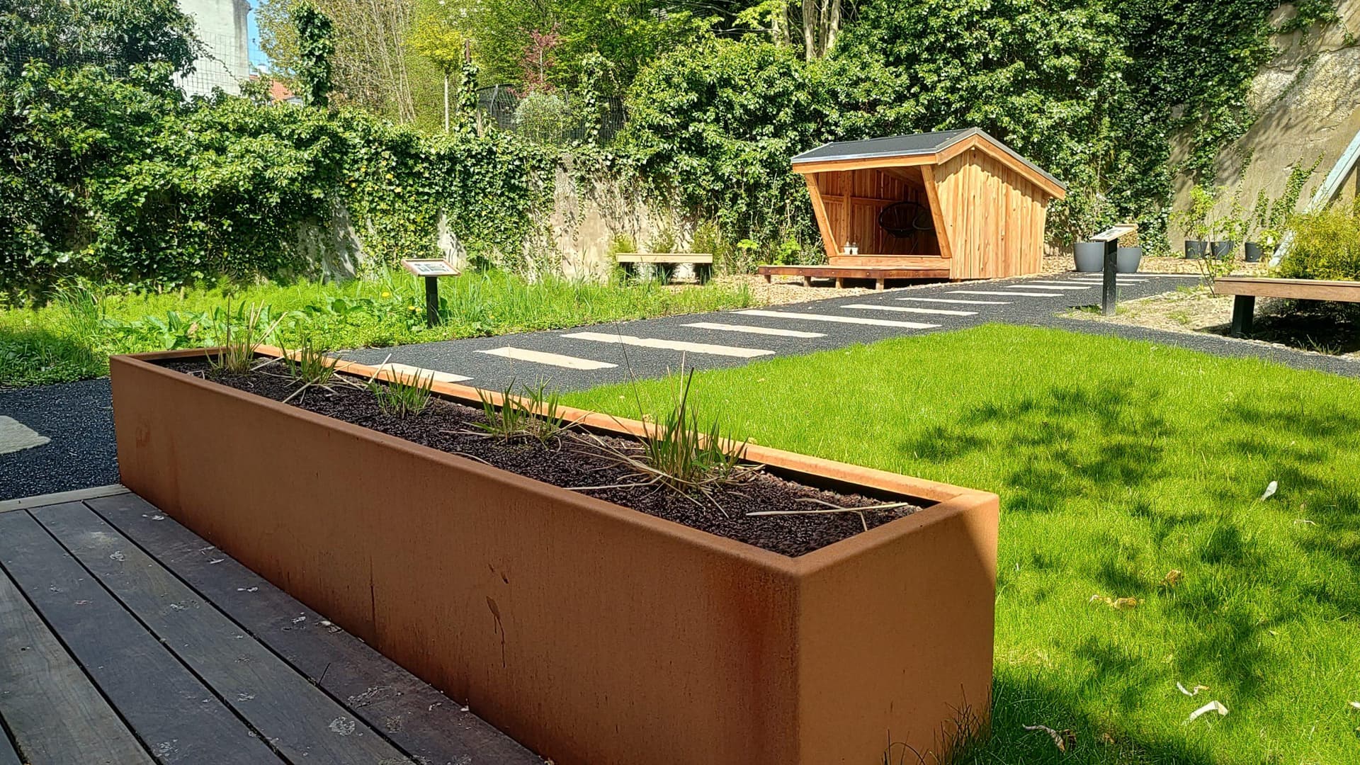Rustfrit stål plantekasse med planter på træ-terrasse foran legehytte og græsplæne