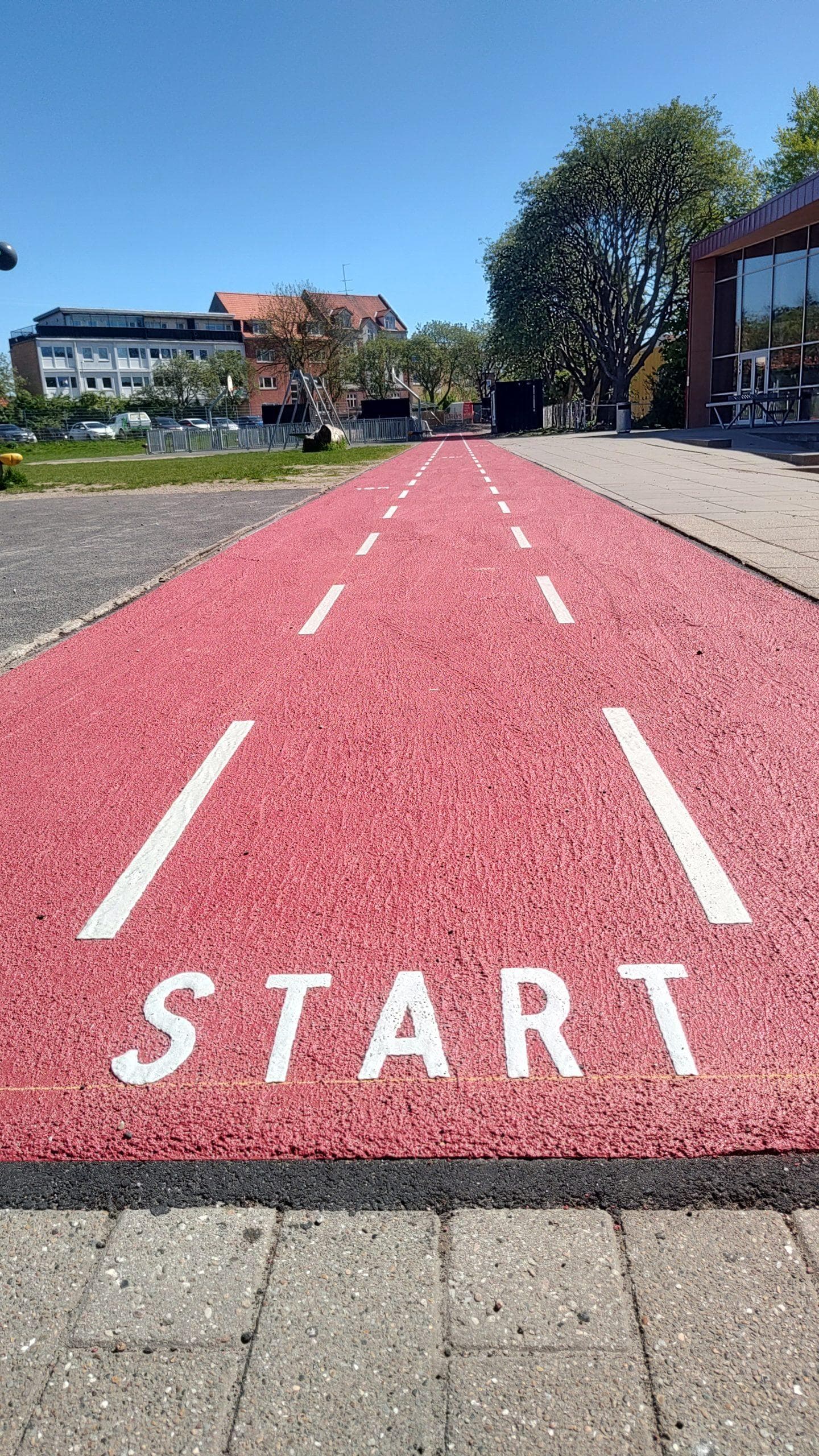 Rødt løbebane med hvide linjer og "START" skrevet på asfalten, omkranset af bygninger og træer
