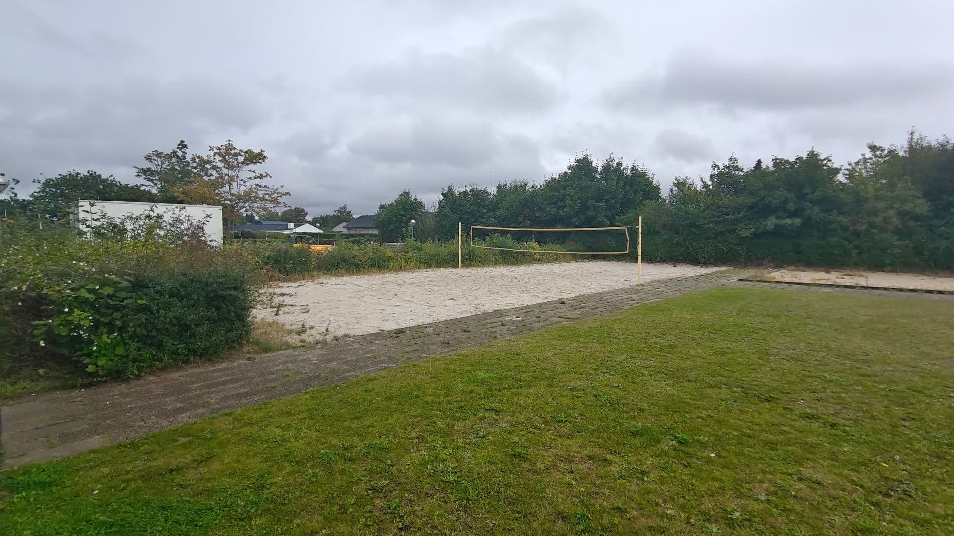 Beachvolleybane med lyst sand og net, omgivet af buske og en græsplæne, inden banen blev nedlagt