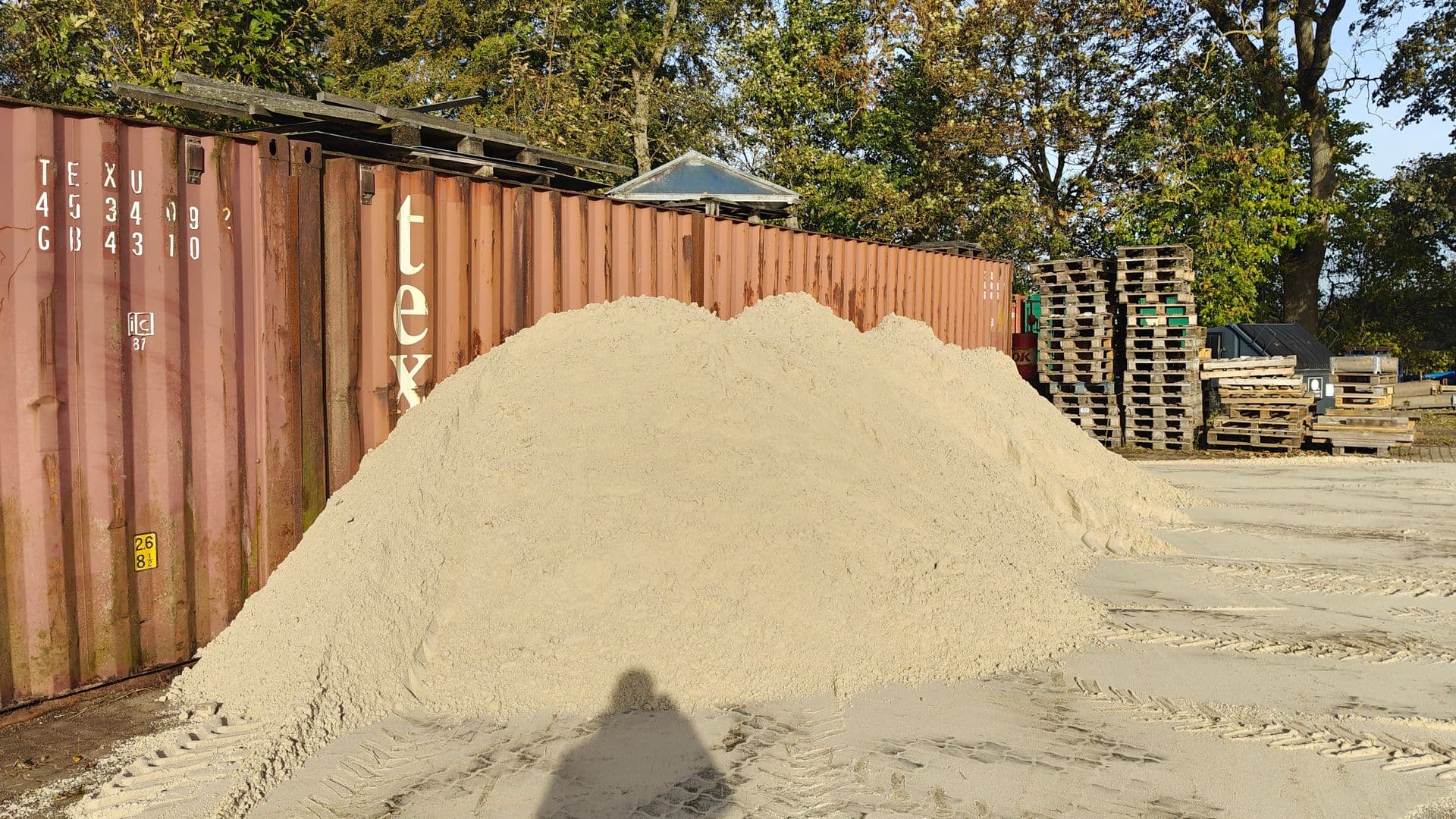 Stor bunke fint, lyst sand foran en container og stablede paller i sollys