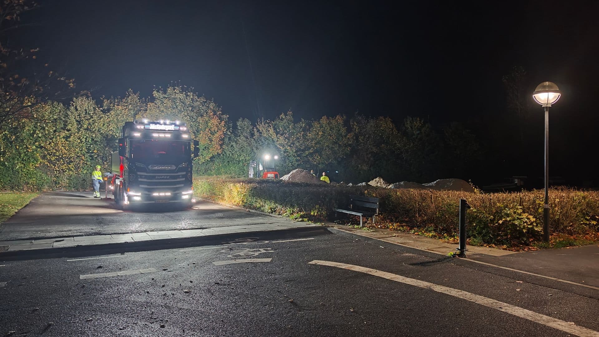 Lastbil og gravemaskine i mørke ved den nedlagte beachbane, hvor sandet ligger i bunker bag en hæk