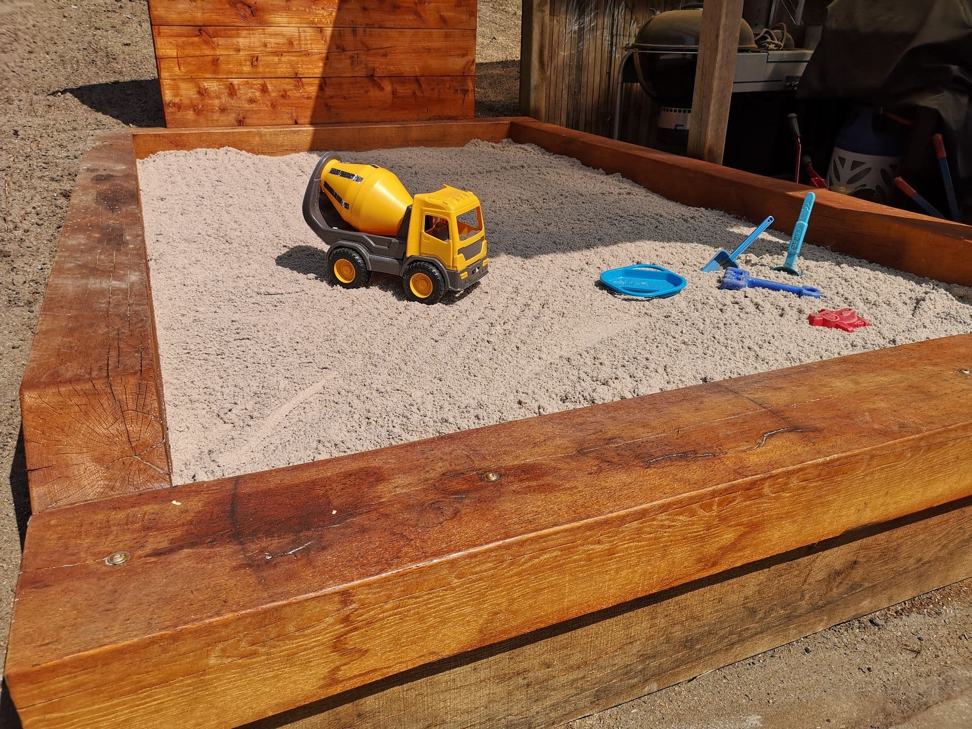 Sandkasse med kant af kraftige træbjælker, fyldt med fint sand og legetøj, blandt andet en gul betonblander